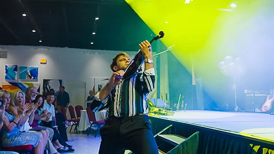 A man playing a violin on a stage in front of an audience, with bright yellow and blue stage lighting and people clapping in the background.