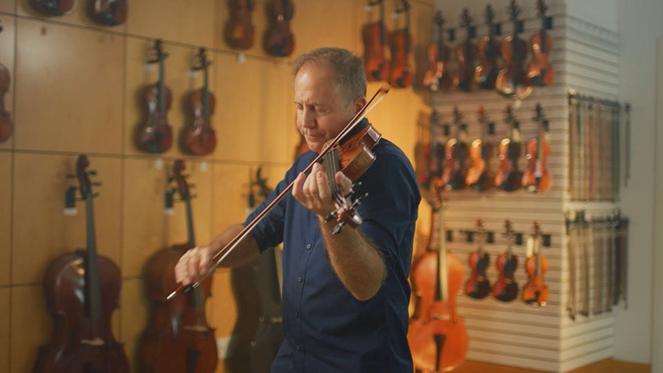 A man in a navy blue shirt playing a violin in a music store, with violins hanging on display on the wooden walls behind him.