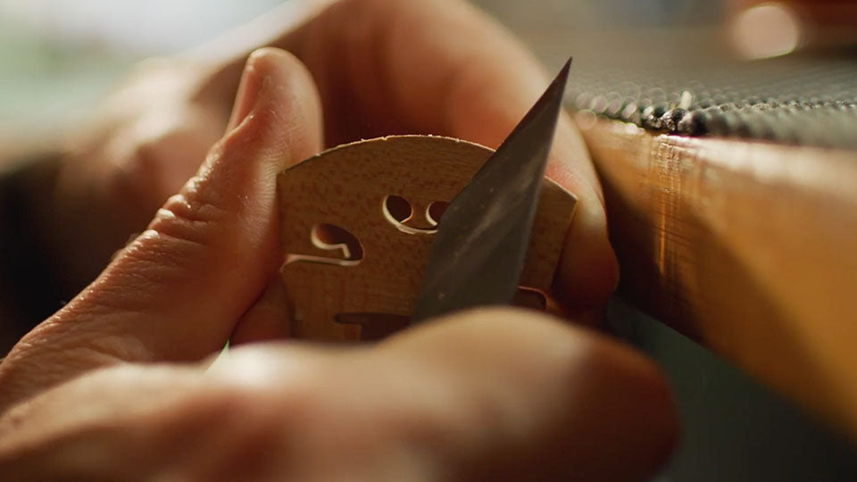 A close-up shot of a person's hands carving a violin bridge with a sharp knife, set against a warm, blurred background.