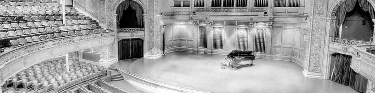 Black-and-white photograph of an elegant concert hall with ornate architecture, rows of seating, and a grand piano on stage