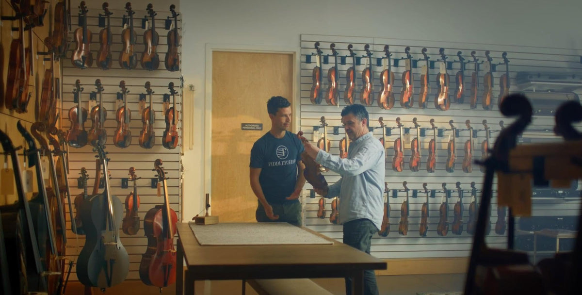 A man in a light blue shirt demonstrates how to hold and position a violin to a customer wearing a dark blue t-shirt inside a violin shop with many violins hanging on the walls.