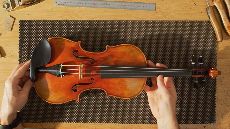 A top-down view of a person holding a wooden violin on a dark textured mat over a wooden workbench, with tools visible around the edges.