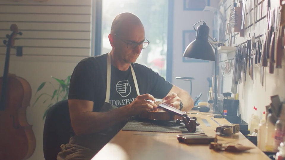 A man wearing glasses and a black t-shirt with a white apron sits at a workbench in a workshop, setting up a student violin. A cello is visible in the background on the left, and tools hang on the wall to the right.