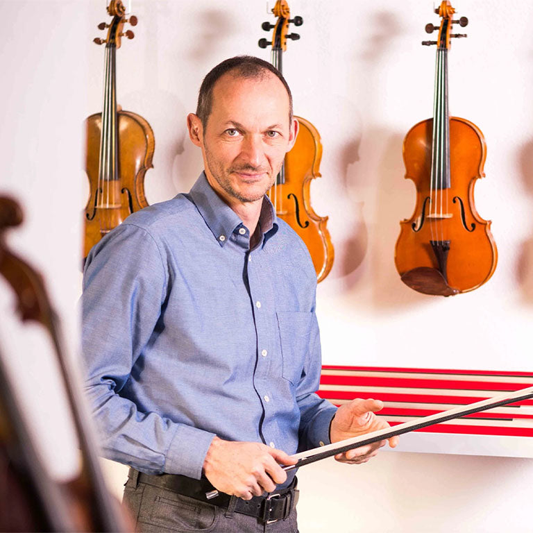 Müsing bow craftsman holding a carbon fiber string instrument bow in his workshop, with violins and violas displayed on the wall behind him.